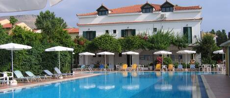 A swimming pool with lounge chairs and umbrellas, surrounded by a building with a tiled roof and greenery.