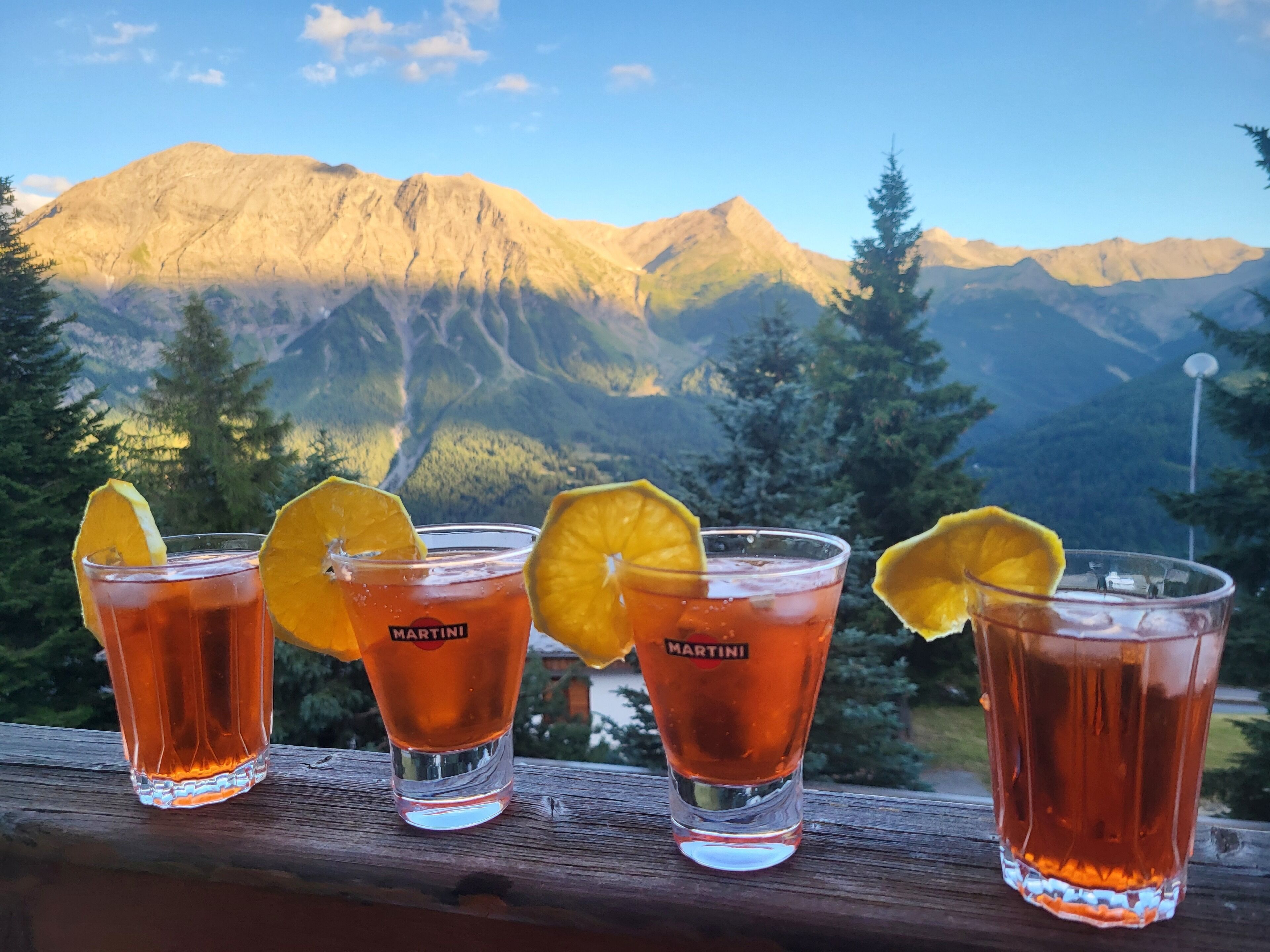 Four glasses of Martini cocktail with orange slices on a wooden railing with a mountain view in the background.