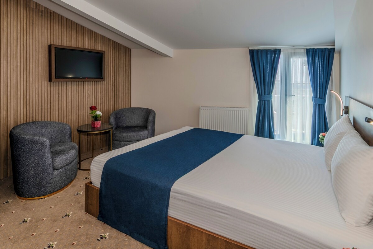 Una habitación de hotel con una cama, dos sillones, un televisor y una ventana con cortinas.