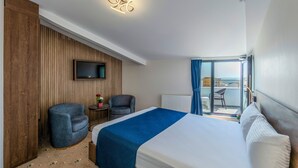 Una habitación de hotel con cama, dos sillones, una mesita con una planta, un televisor montado en la pared y un balcón con mesa y sillas.