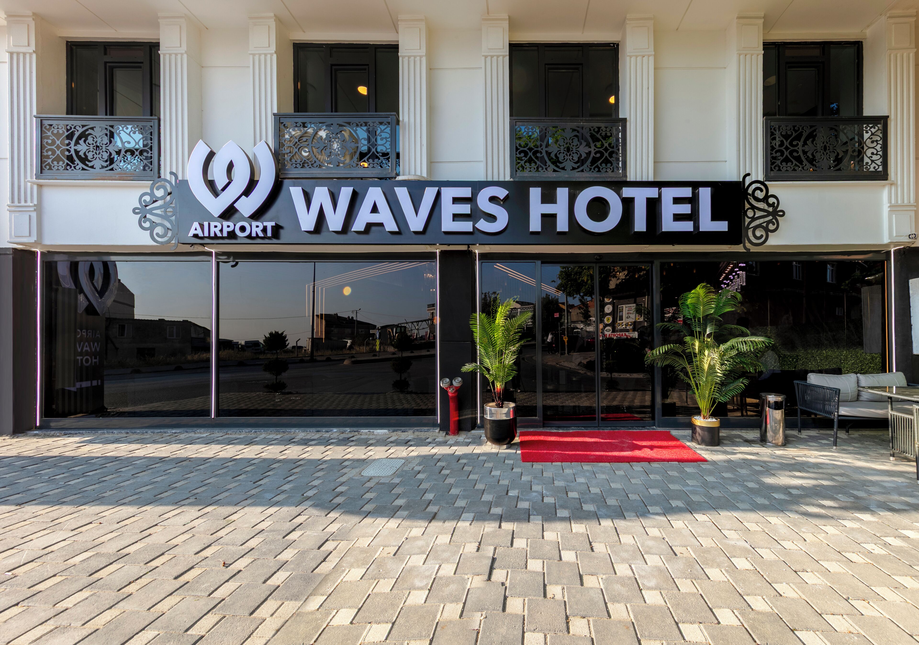 Exterior del Waves Hotel, con alfombra roja, plantas en macetas y un diseño arquitectónico moderno.