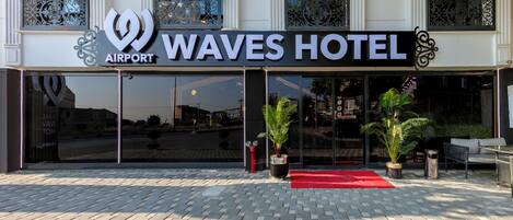 Exterior del Waves Hotel, con alfombra roja, plantas en macetas y un diseño arquitectónico moderno.