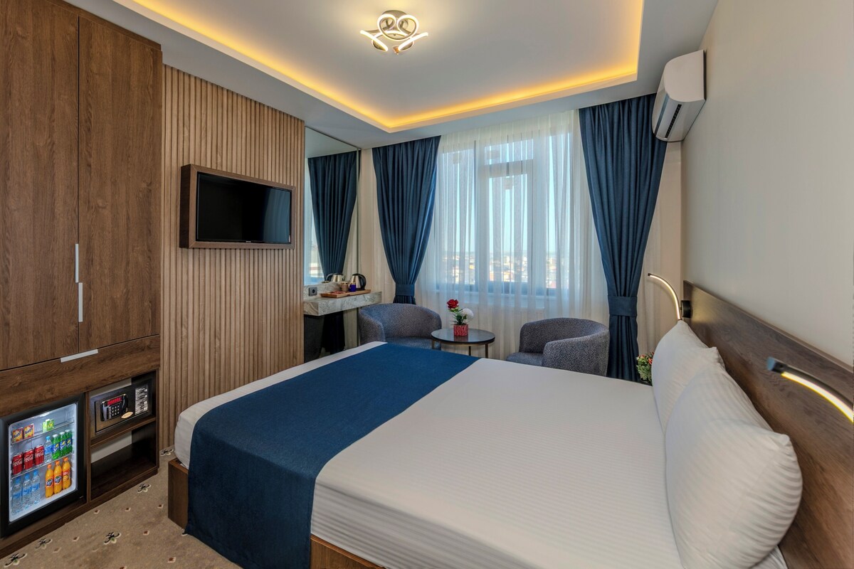 Habitación de hotel con una cama grande, un televisor montado en la pared, un mini bar y un área de estar con sillas y una mesa.