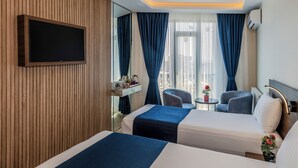 Habitación de hotel con dos camas, un televisor de pantalla plana montado en la pared, una mesita con un florero y una ventana con cortinas azules.