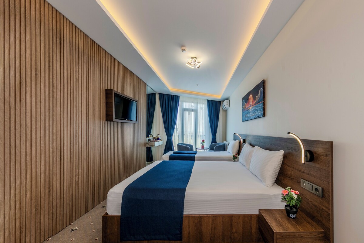 Una habitación de hotel moderna con una cama grande, una mesita de noche con lámpara, un televisor montado en la pared y una ventana con cortinas.