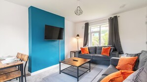 A modern living room with a blue accent wall, a flat-screen TV, a sofa with orange cushions, and a coffee table. - Kneeton Vale (Nottingham)
