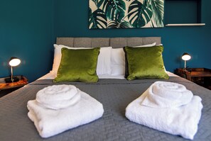 Una cama bien hecha con dos almohadas verdes y dos blancas, una colcha gris y dos toallas blancas enrolladas sobre una colcha gris con textura.