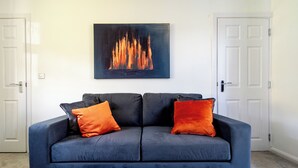 A dark blue sofa with two orange cushions, a painting of a fire on the wall, and white doors on either side. - Kneeton Vale (Nottingham)