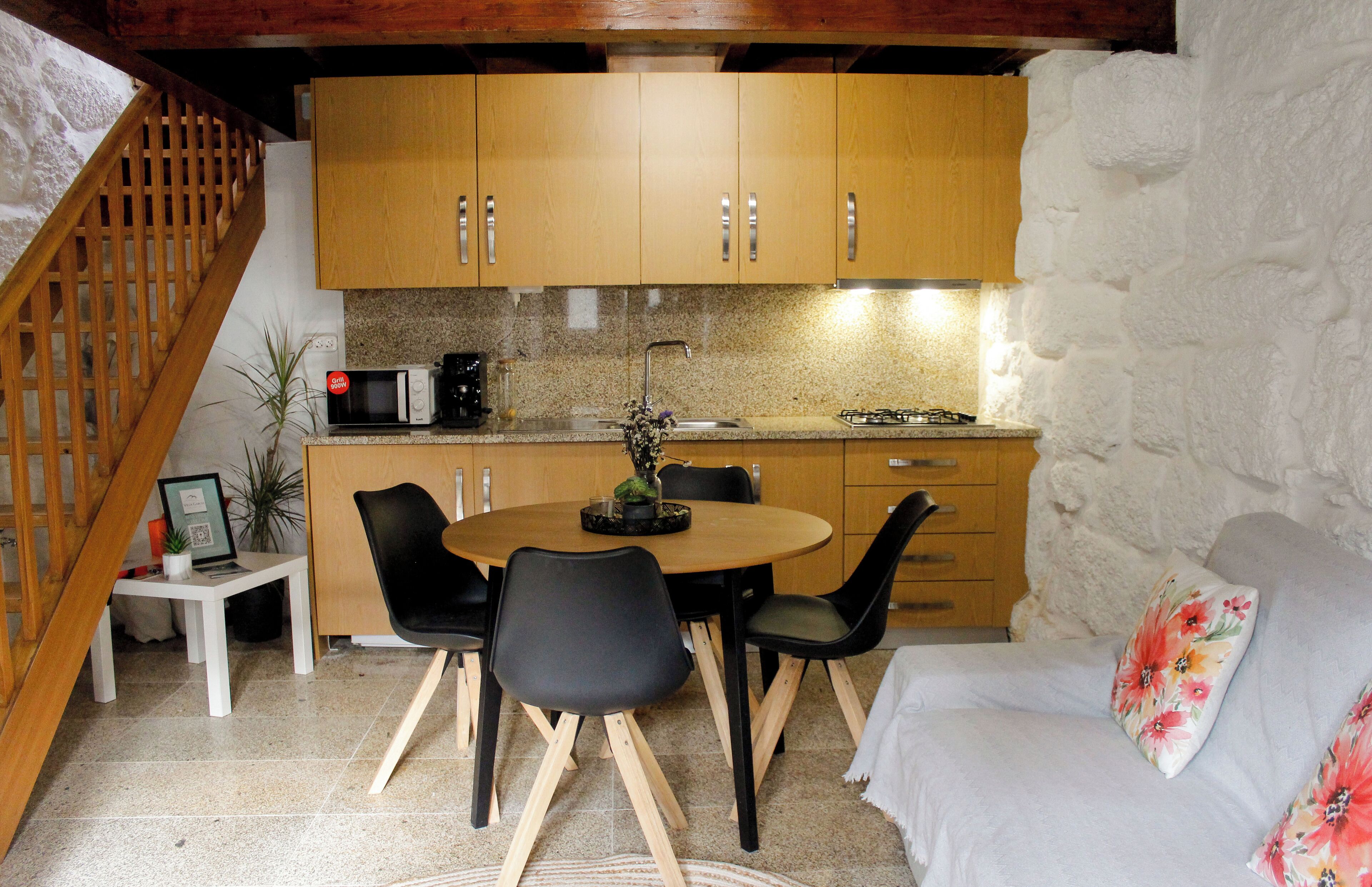 A cozy kitchen with wooden cabinets, a dining table with chairs, a small white table with a laptop, and a sofa with a floral pillow.