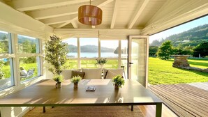 A sunlit room with a wooden table, potted plants, and a view of a lake and greenery. - The Cliffs - Cedeira Bay (Cedeira)