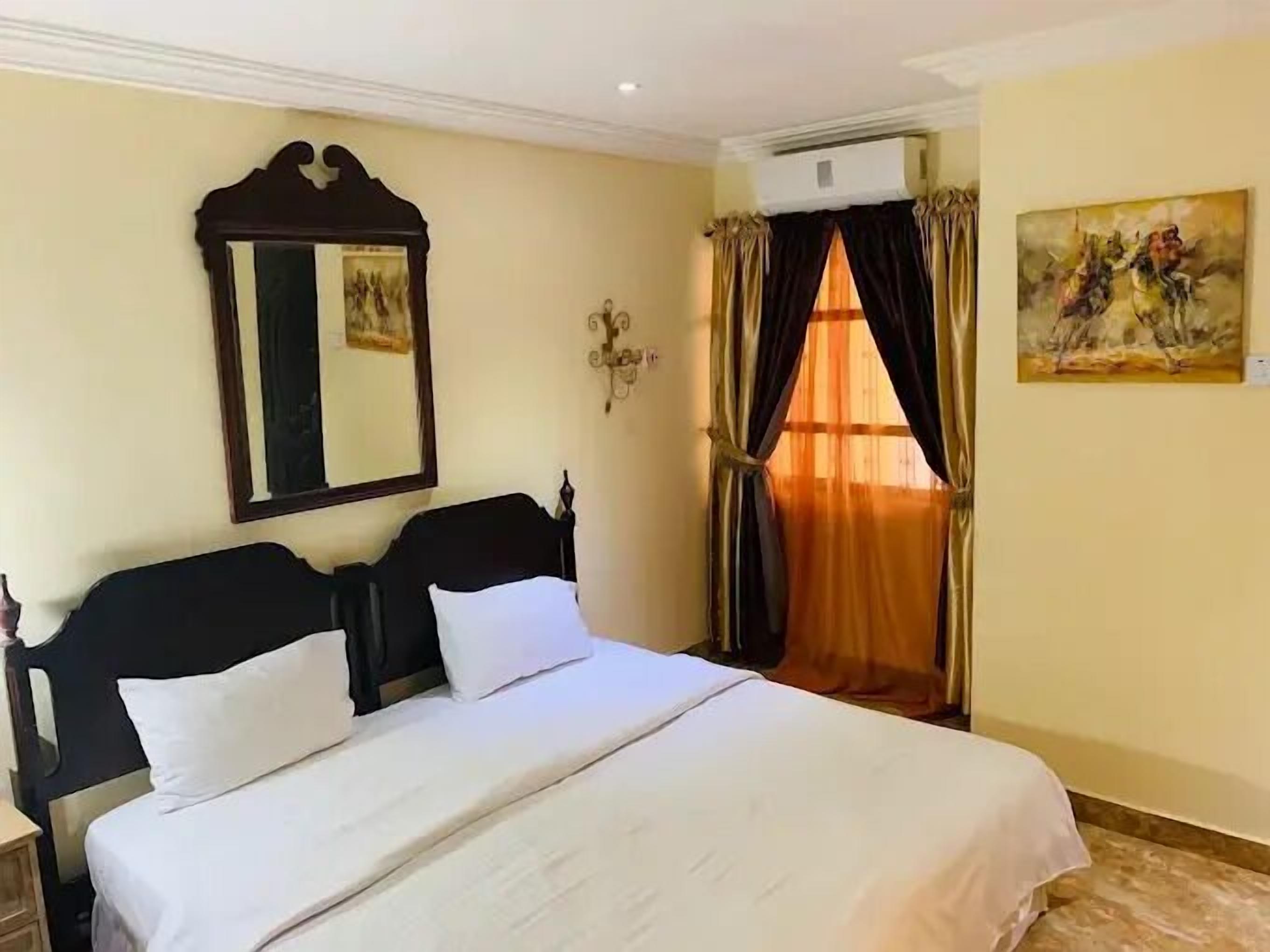 Une chambre d’hôtel avec un lit, une table de chevet, un climatiseur fixé au mur, un tableau et une fenêtre avec des rideaux.