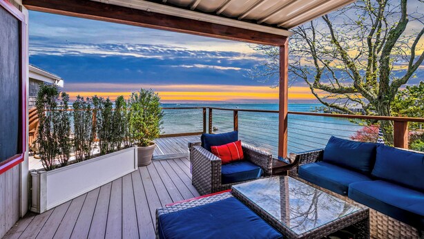 Una terrazza coperta con mobili in vimini, un tavolo in vetro e vista sull'oceano al tramonto.