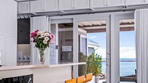 Una cucina moderna con bancone e sgabelli in legno, un camino e vista mare attraverso ampie porte scorrevoli.