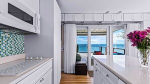 Una cucina moderna con mobili bianchi, schienale piastrellato e vista sull'oceano tramite una porta in vetro.