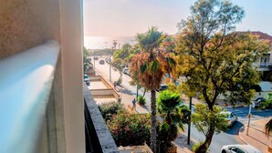 A view from a window overlooking a street with palm trees, parked cars, and a building with a red roof. - Herzlia Pituah apartment with Pool, by the Acadia Beach! (Herzliya)