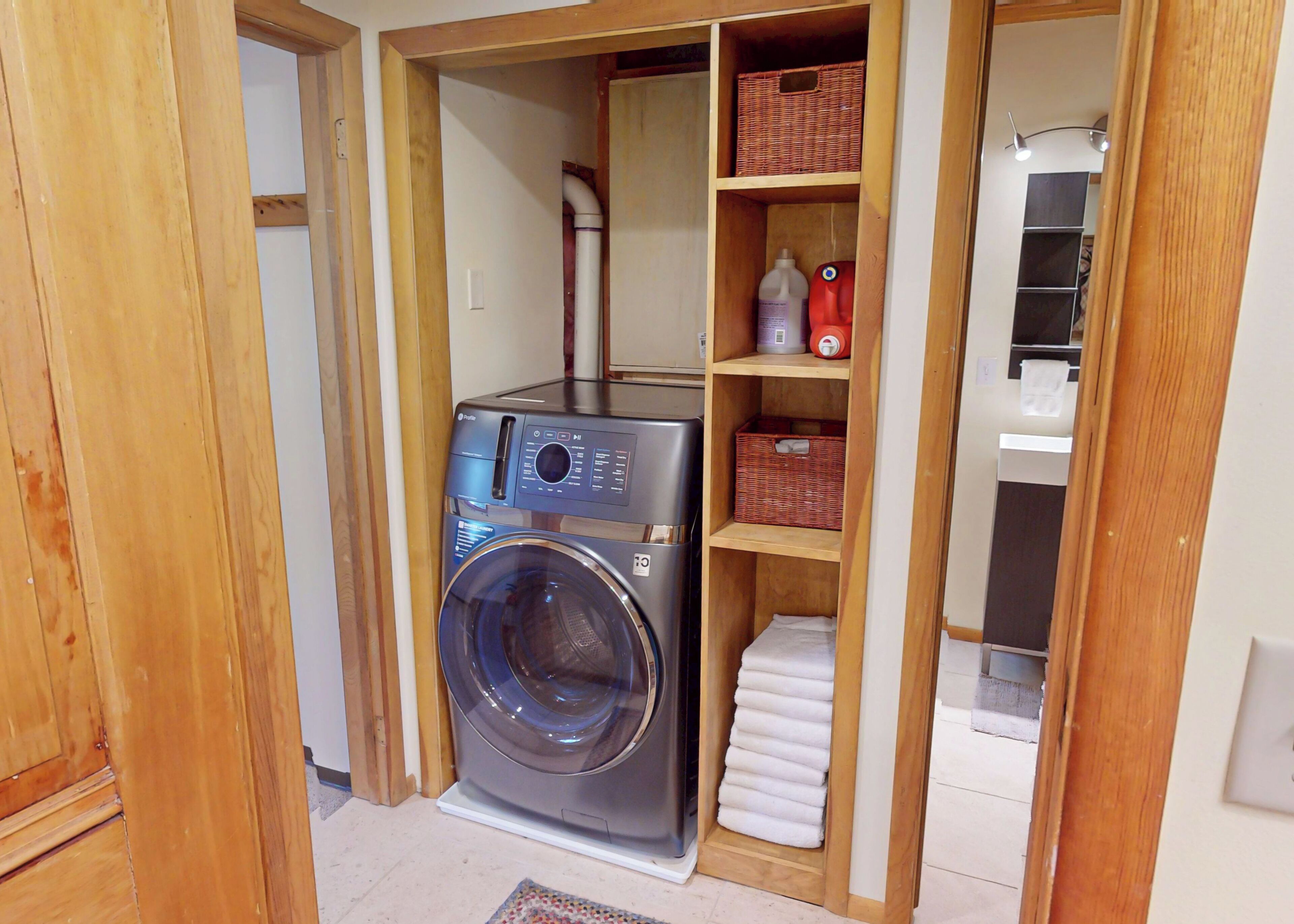 A compact laundry room with a front-loading washing machine, wooden shelving with baskets, and a small drying rack.