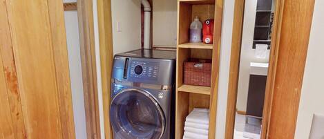 A compact laundry room with a front-loading washing machine, wooden shelving with baskets, and a small drying rack.