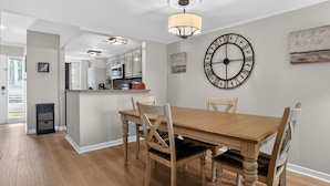 Un comedor con una mesa y sillas de madera, un reloj de pared y una cocina al fondo.