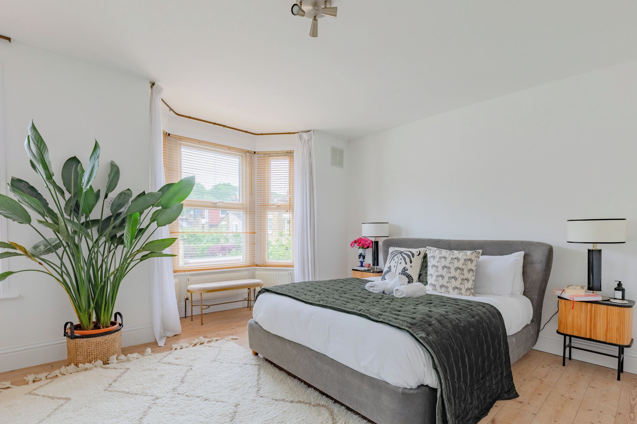 A bedroom with a bed, bedside table, lamp, potted plant, and a window with curtains.