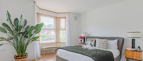 A bedroom with a bed, bedside table, lamp, potted plant, and a window with curtains.