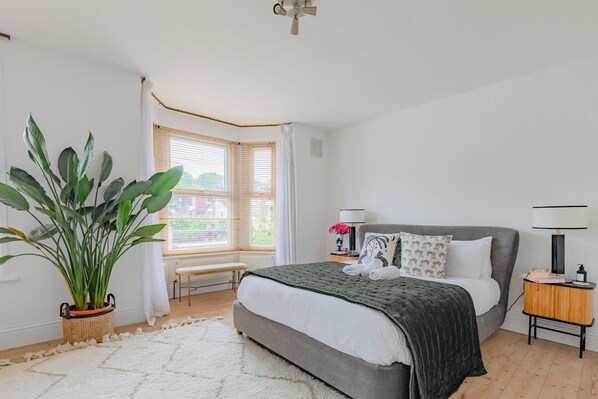 A bedroom with a bed, bedside table, lamp, potted plant, and a window with curtains.