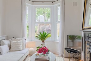 A room with a white sofa, a round coffee table with books and a plant, a TV on a stand, and a bay window with curtains.