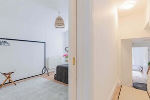 A hallway with a white wall, a radiator, a grey rug, a wooden stool, a hanging light fixture, and a door leading to another room.