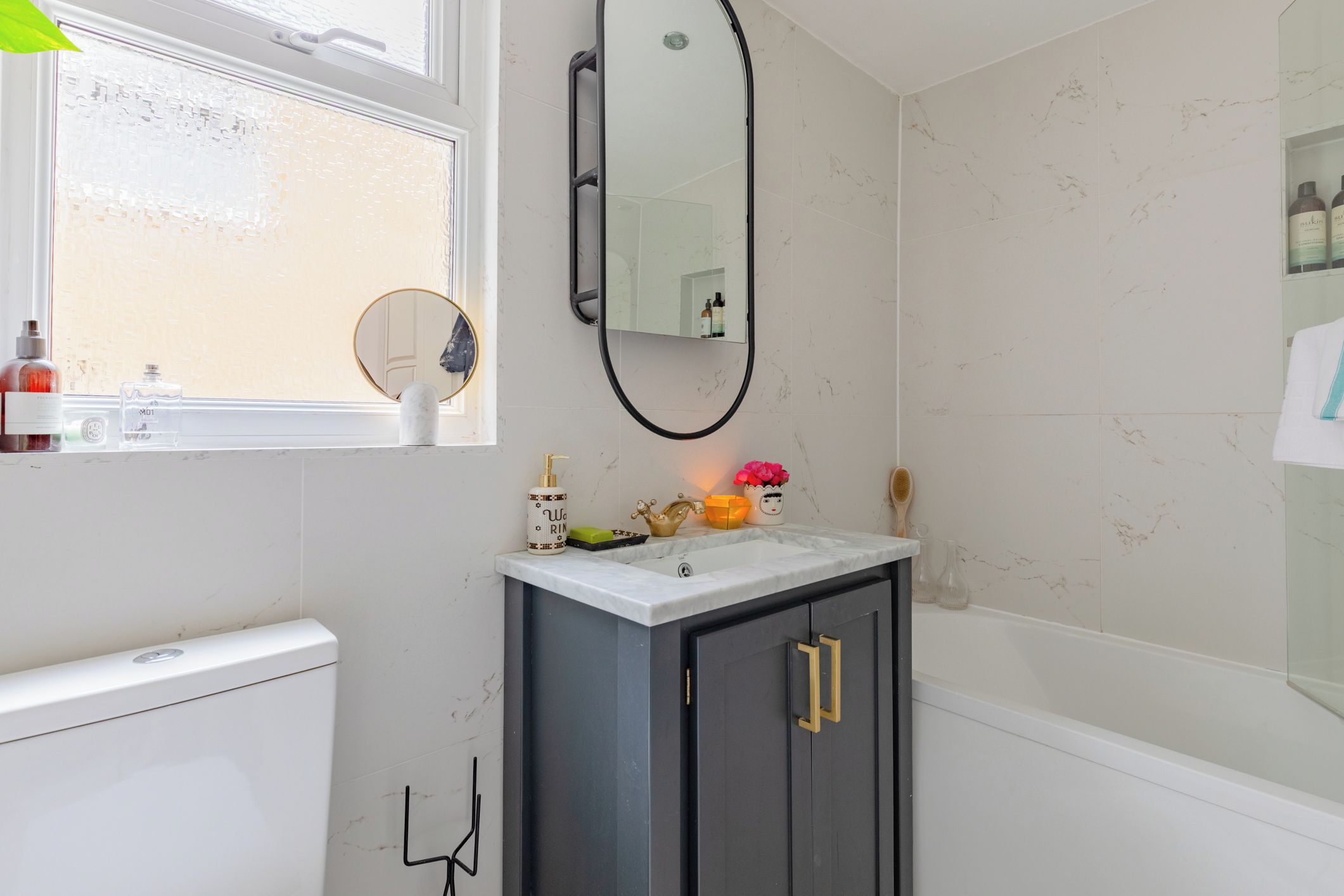 A bathroom with a white toilet, a sink with a marble countertop, a mirror, and toiletries on the windowsill.