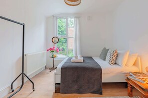 A bedroom with a bed, a bedside table with books, a window with curtains, and a radiator. - Luxuriously Curated 4BD Home, Stoke Newington (Greater London)