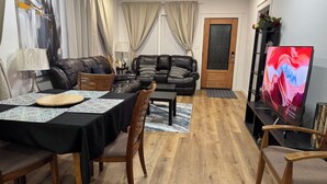 A living room with a dining area, featuring a black leather sofa, a wooden dining table, and a TV on a stand.