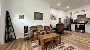 Sala de estar con sofá, mesa de centro y alfombra estampada. Cocina con armarios y estufa. Una escalera y objetos decorativos a la izquierda.
