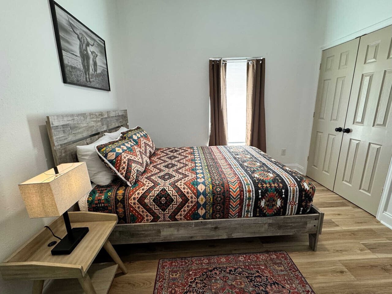 Ein Schlafzimmer mit einem hölzernen Bett, gemusterten Betttextilien, einer Nachttischlampe, einem Teppich und einer Tür mit einem Spiegel.