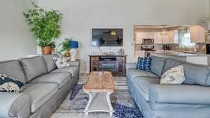A modern living room with two grey sofas, a wooden coffee table, and a flat-screen TV. There is a kitchen area with white cabinets and a microwave.
