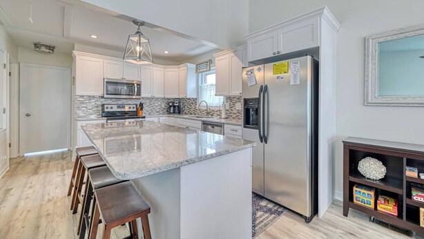 A modern kitchen with white cabinets, a large island with bar stools, stainless steel appliances, and a hanging light fixture.
