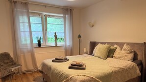 Un dormitorio con una cama grande, una ventana con cortinas, un radiador, una lámpara de noche y una pequeña mesa con una silla.
