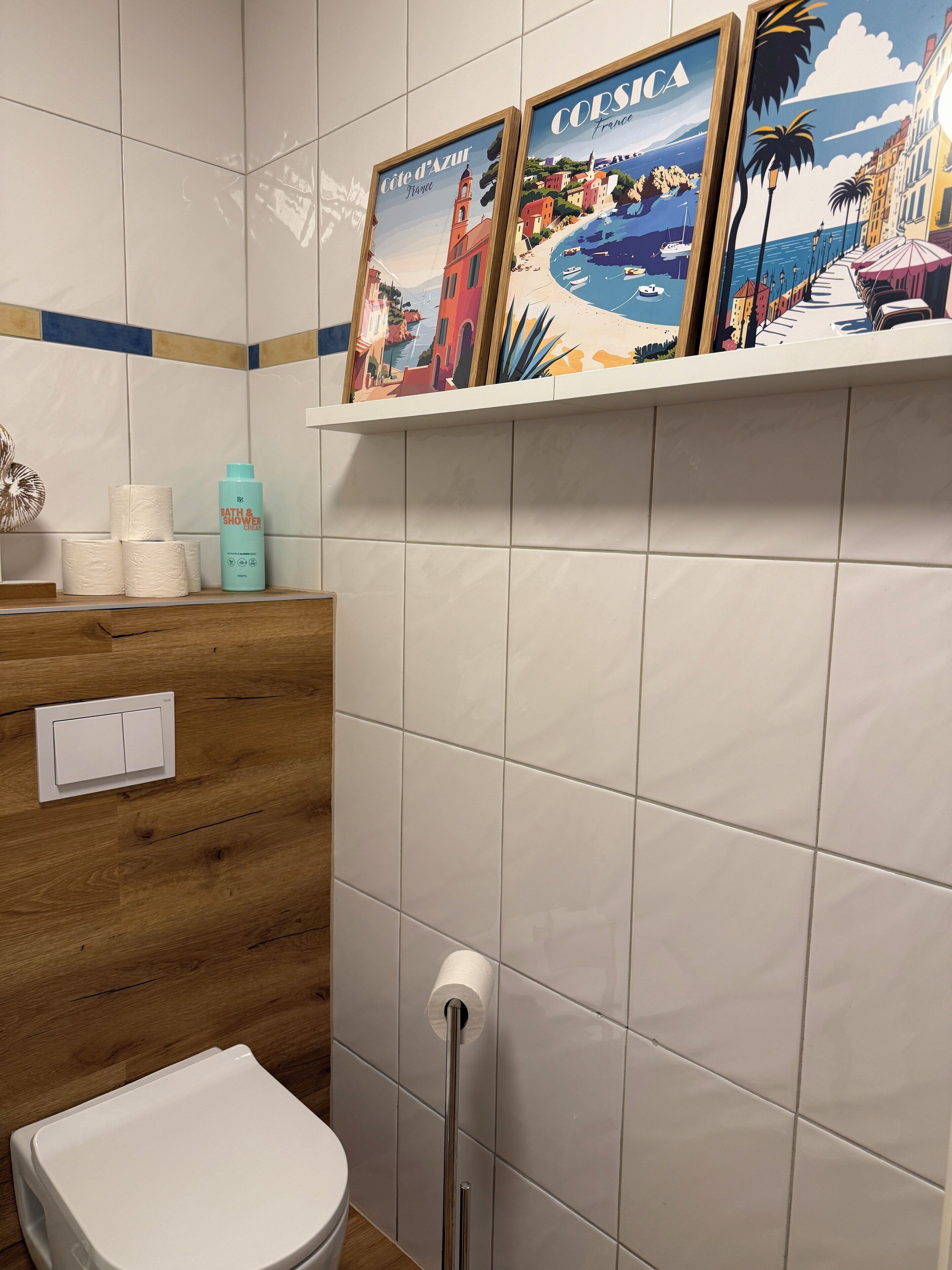 Un baño con tocador de madera, inodoro y estante con fotografías enmarcadas de paisajes costeros.