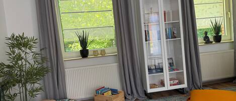 A room with a large window, a white bookshelf, a small wooden toy car, a pink ottoman, and a patterned rug.
