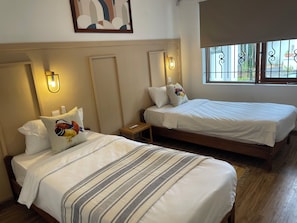 A hotel room with two beds, each with white bedding and decorative pillows, a small bedside table, and wall-mounted light fixtures.