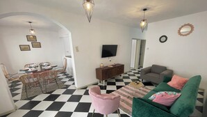 A living room with a checkered floor, a green sofa, a pink chair, a dining area with a table and chairs, and a television on the wall. - Frida, 3-bedroom apartment just a few minutes' walk from Blue Bay lagoon. (Blue Bay)