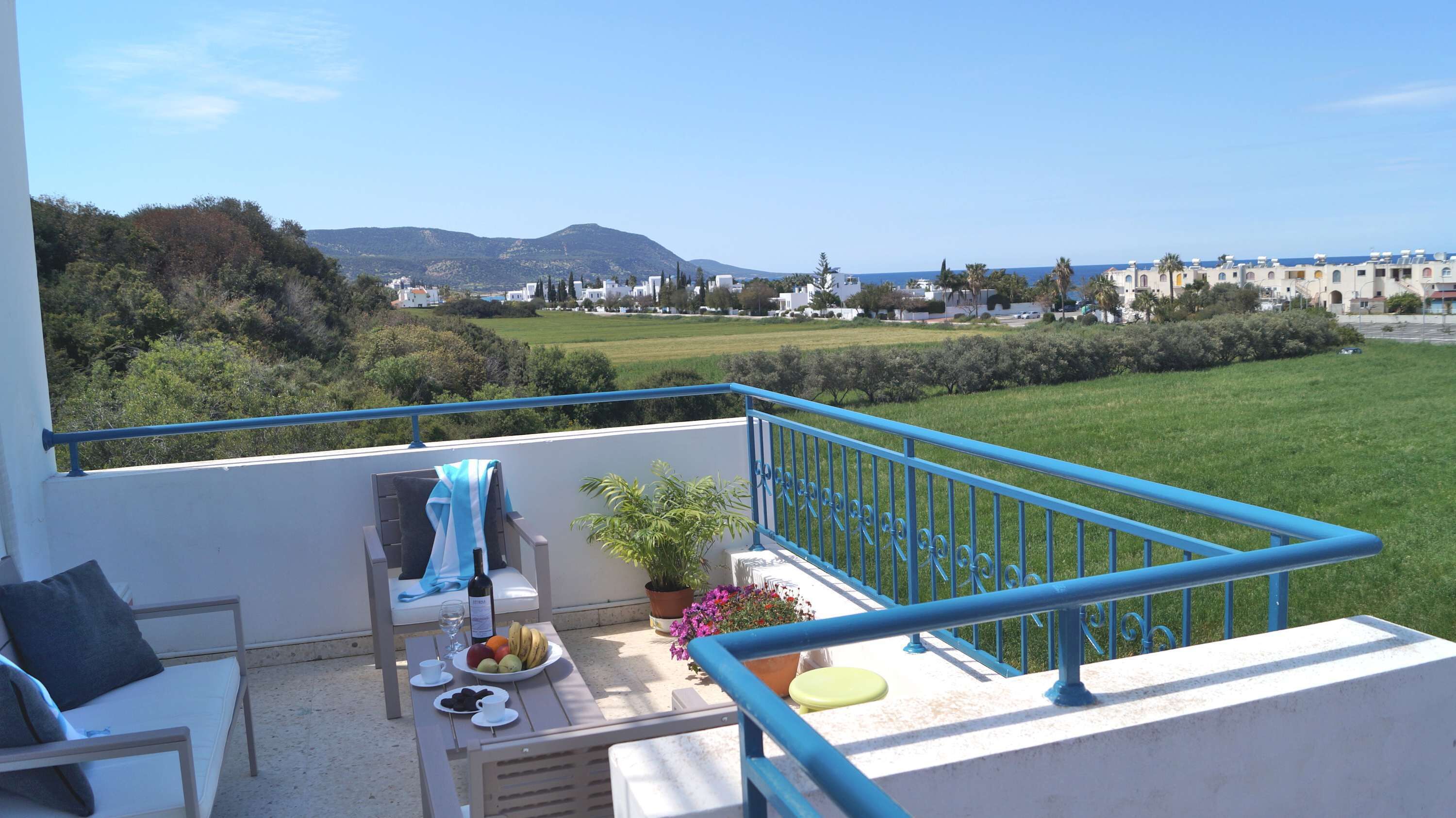 Un balcon avec vue sur la mer et les collines, une table avec de la nourriture et des boissons, et une chaise avec une serviette.