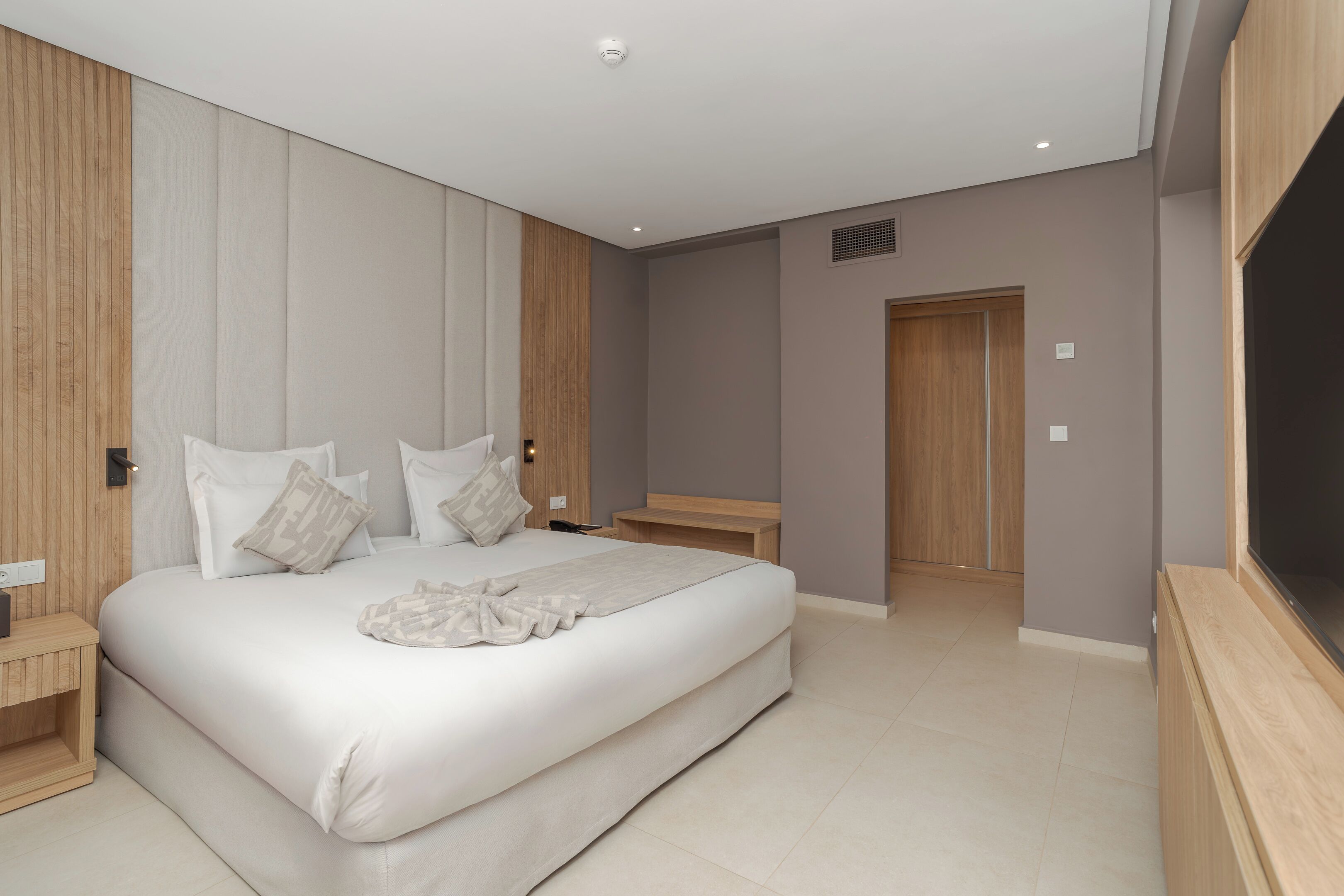 Un dormitorio moderno con una cama grande, paneles de madera, una mesita de noche y una televisión empotrada.