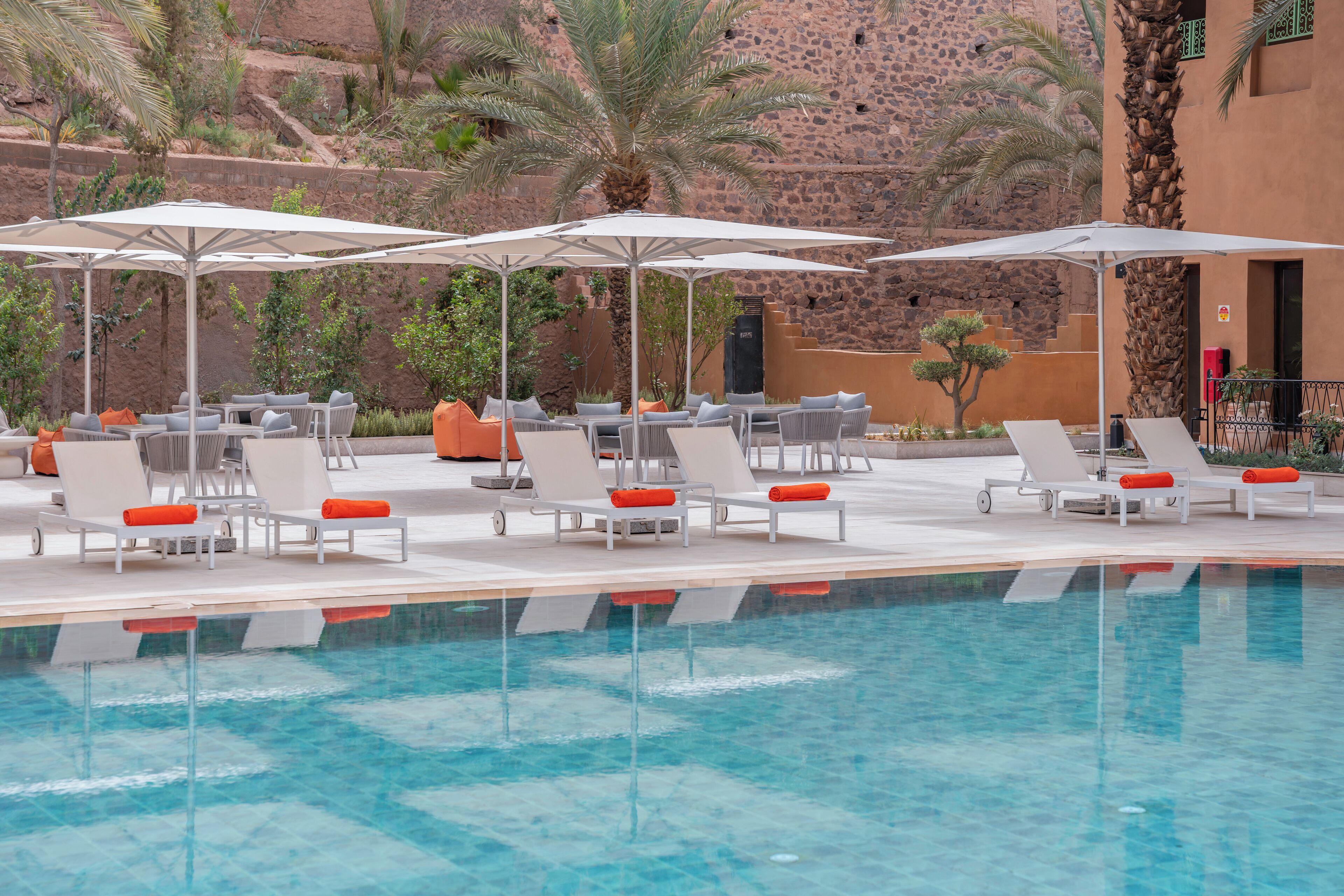 Un área de piscina con sillones de descanso y sombrillas, rodeada de palmeras y un muro de piedra.