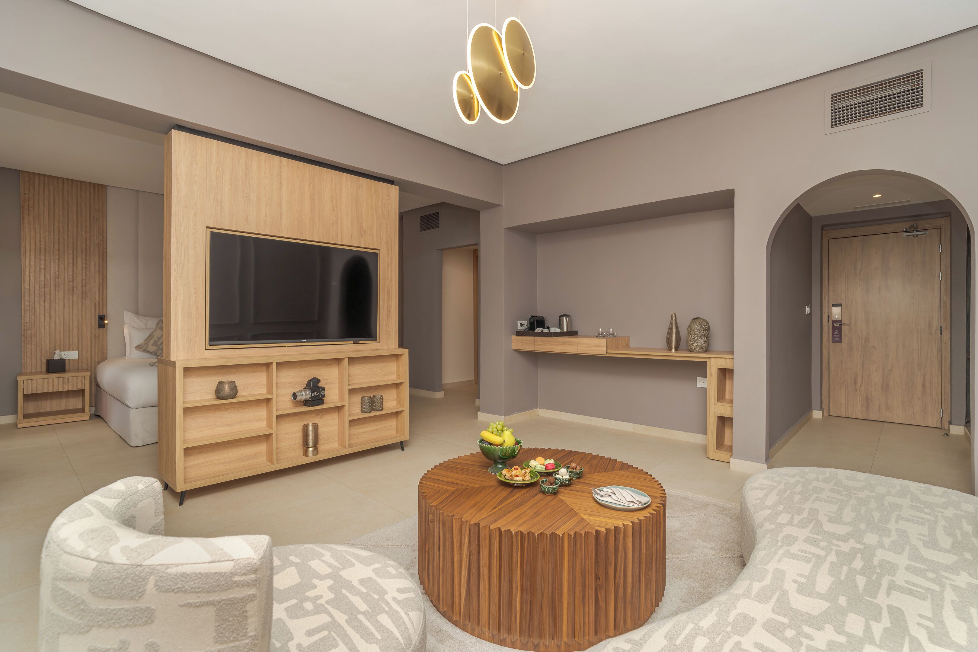 A modern living room with a large flat-screen TV, wooden furniture, a round coffee table with fruits, and a patterned sofa.