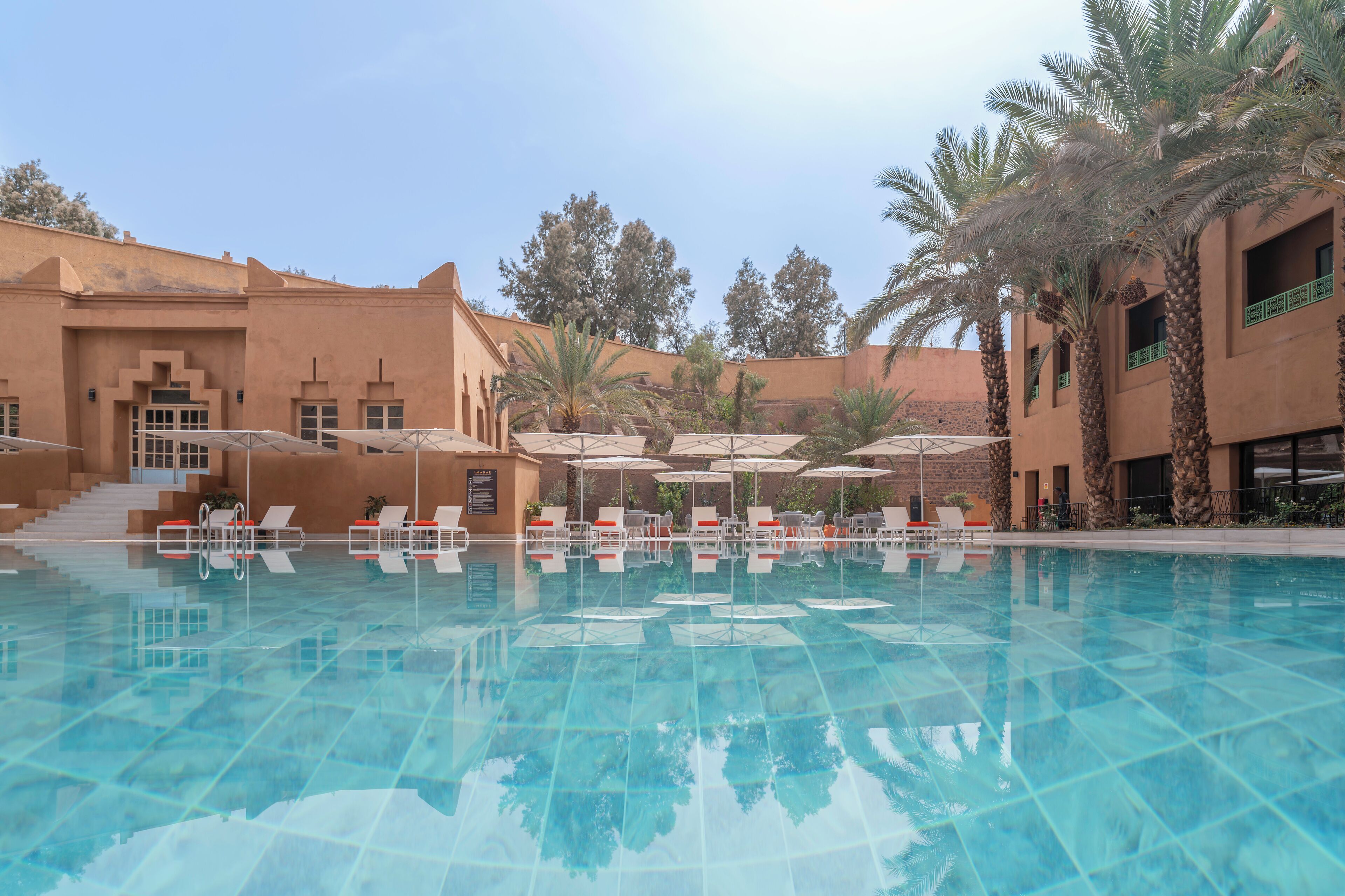 A swimming pool with lounge chairs and umbrellas, surrounded by palm trees and a building with a terracotta facade.