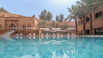 A swimming pool with lounge chairs and umbrellas, surrounded by palm trees and a building with a terracotta facade.