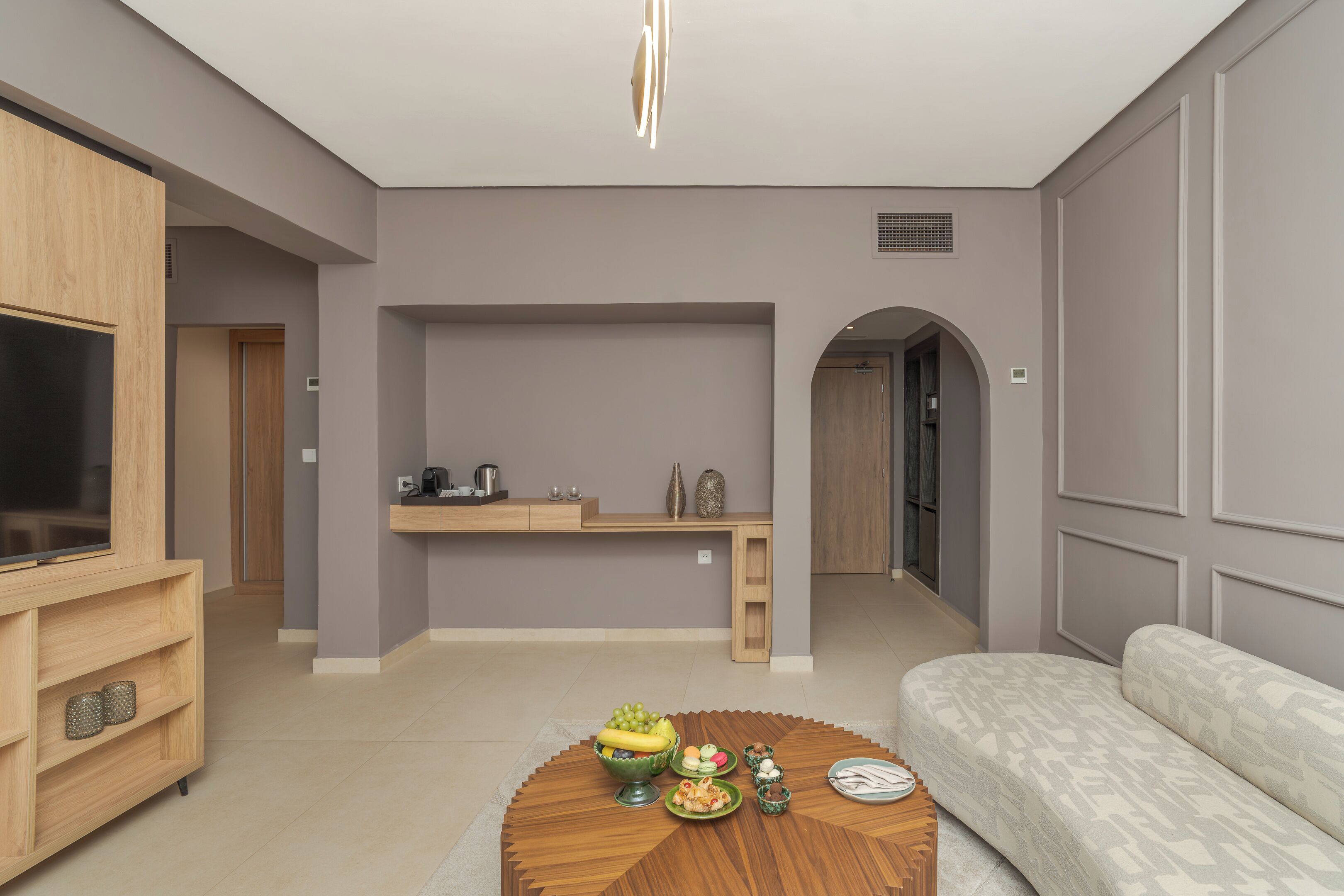 A modern living room with a sofa, a coffee table with fruits, a TV, and a kitchen area in the background.