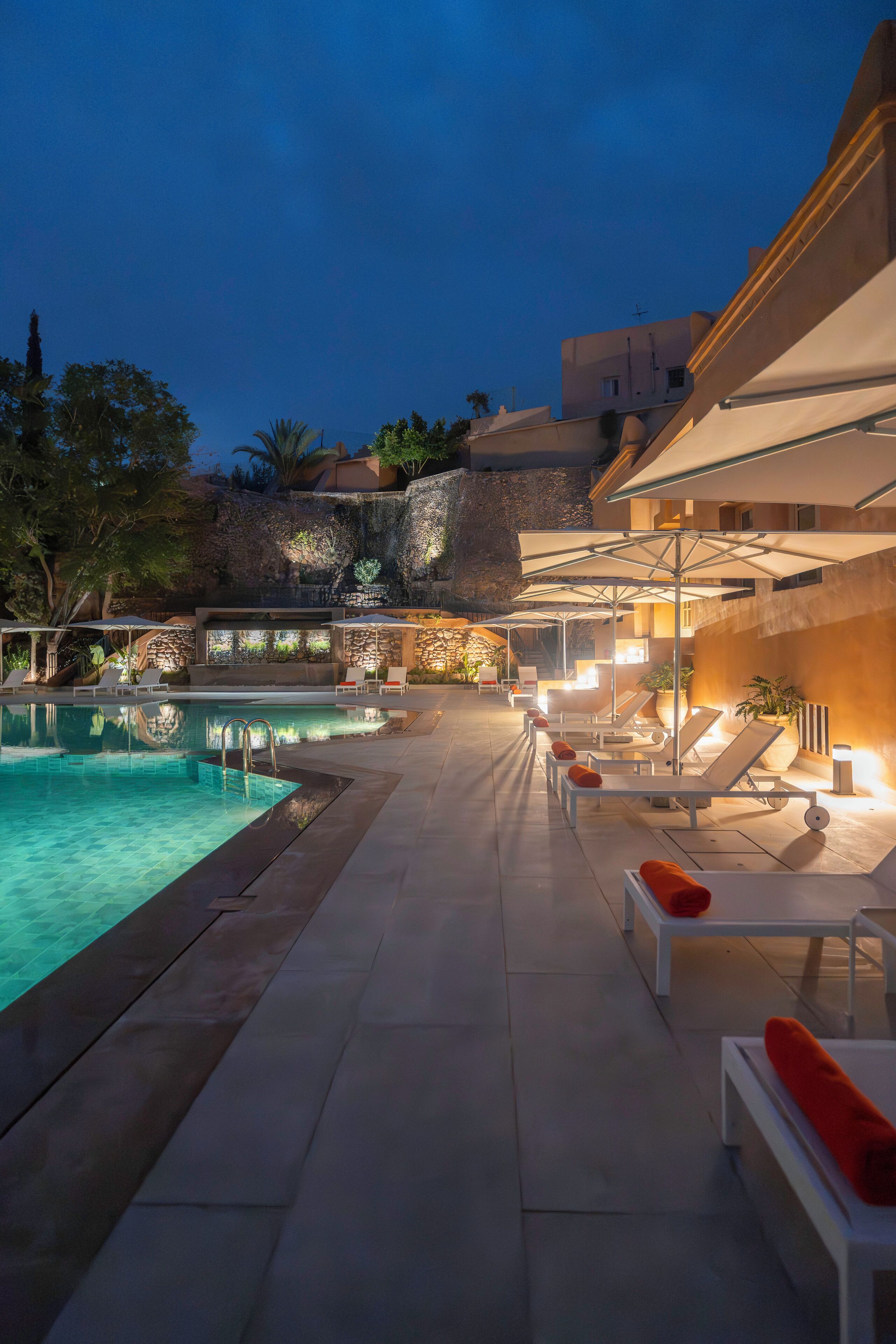 Una piscina exterior con sillas de descanso y sombrillas, rodeada de muros de piedra y vegetación.