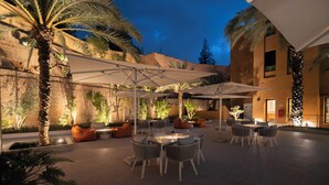 An outdoor patio area with tables and chairs, palm trees, and a building in the background. - AMANAR Ouarzazate Boutique Hôtel & SPA  (Ouarzazate)