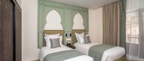 Une chambre d’hôtel avec deux lits, une porte en bois, une tête de lit verte et une fenêtre avec des rideaux.