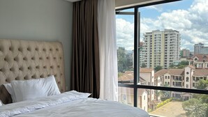Une chambre moderne avec un grand lit, une tête de lit capitonnée et une vue sur les immeubles de la ville par une grande fenêtre.
